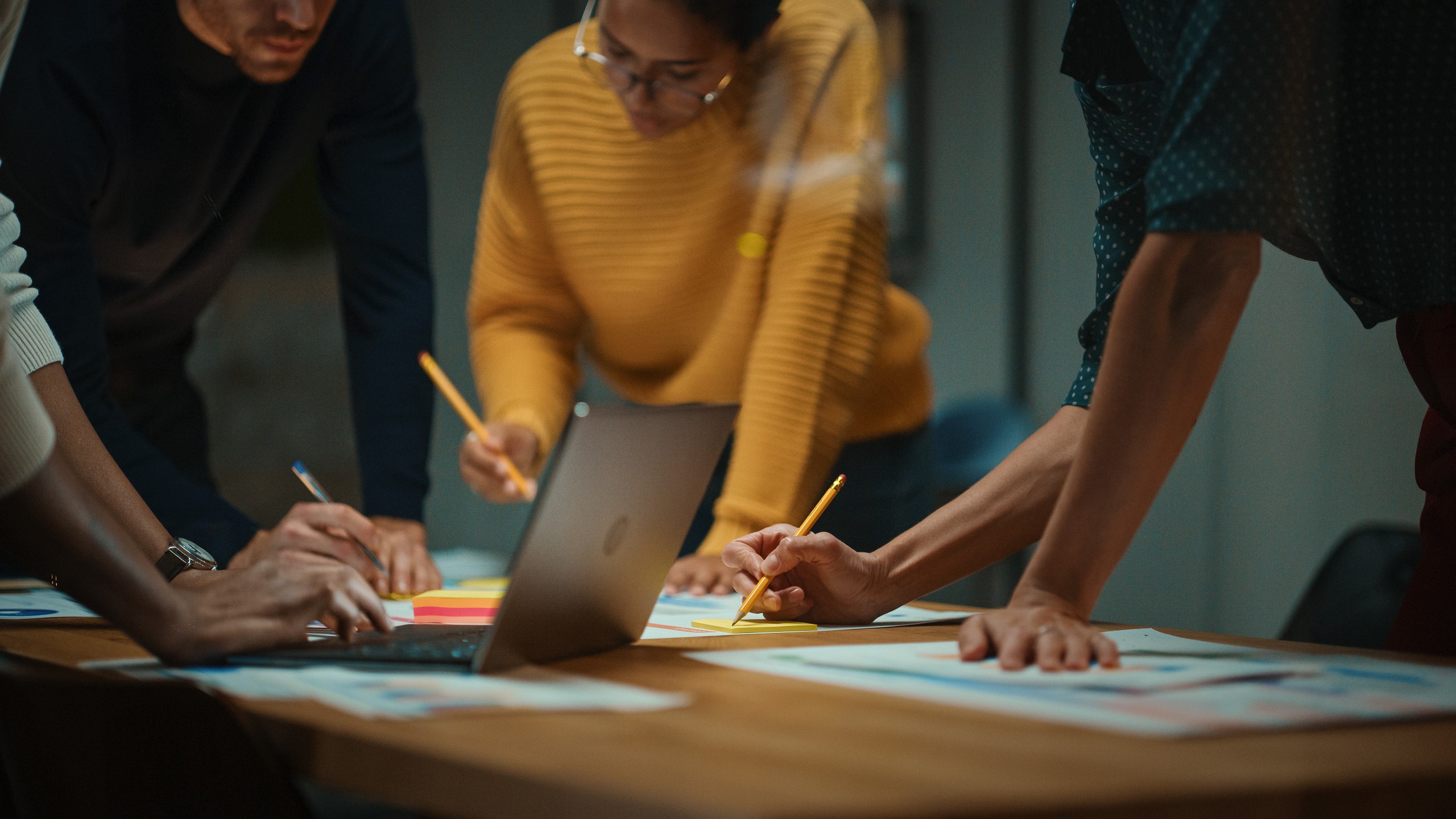 A table with laptop in the middle. People surround the table focusing on a sheet of paper with pencils in their hands.