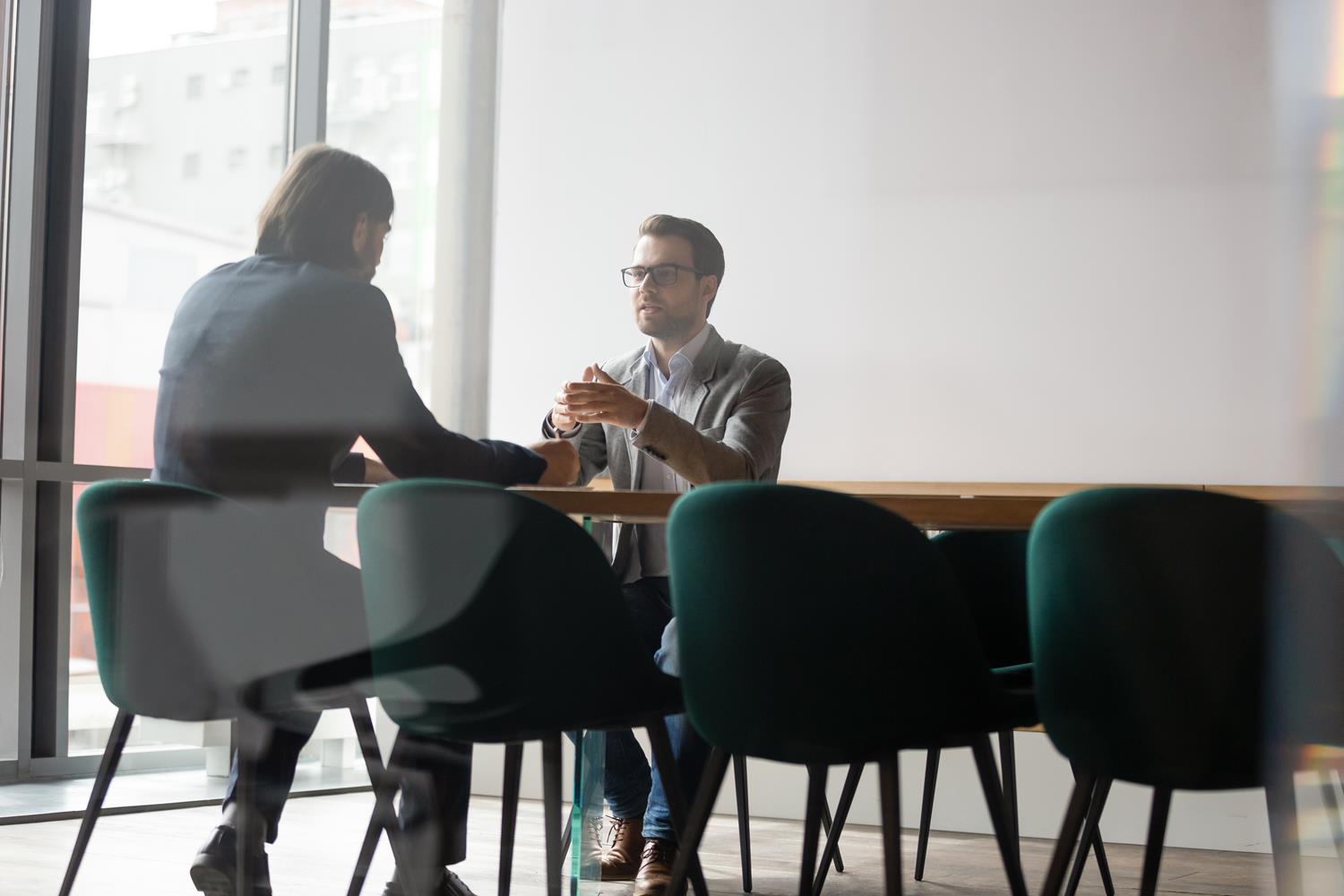 Two men sat at a table in conversation. they wear suits.
