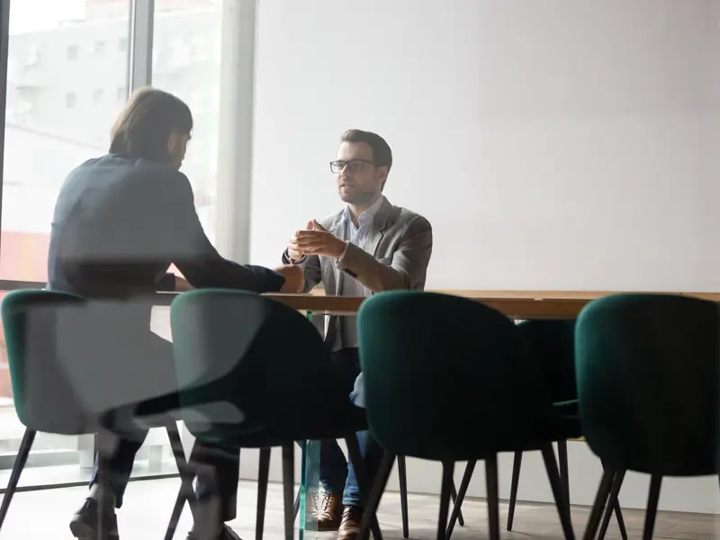 Two men sat at a table in conversation. they wear suits.