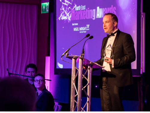 Our Director, Chris Lucarelli in a black suit and bow. He is standing on stage at the Northern Marketing Awards talking into a microphone. He holds an award. 