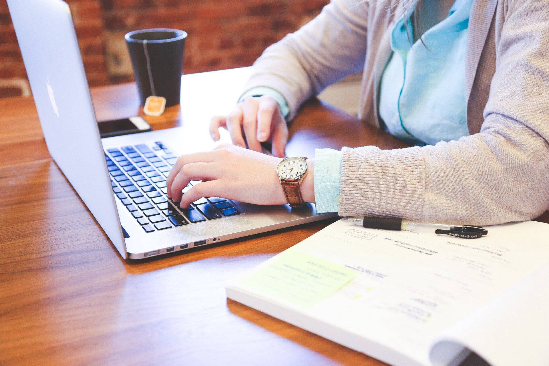Person seated at a work station working on a laptop with an open book to the left of the person and a cup of tea with cellphone on the right.