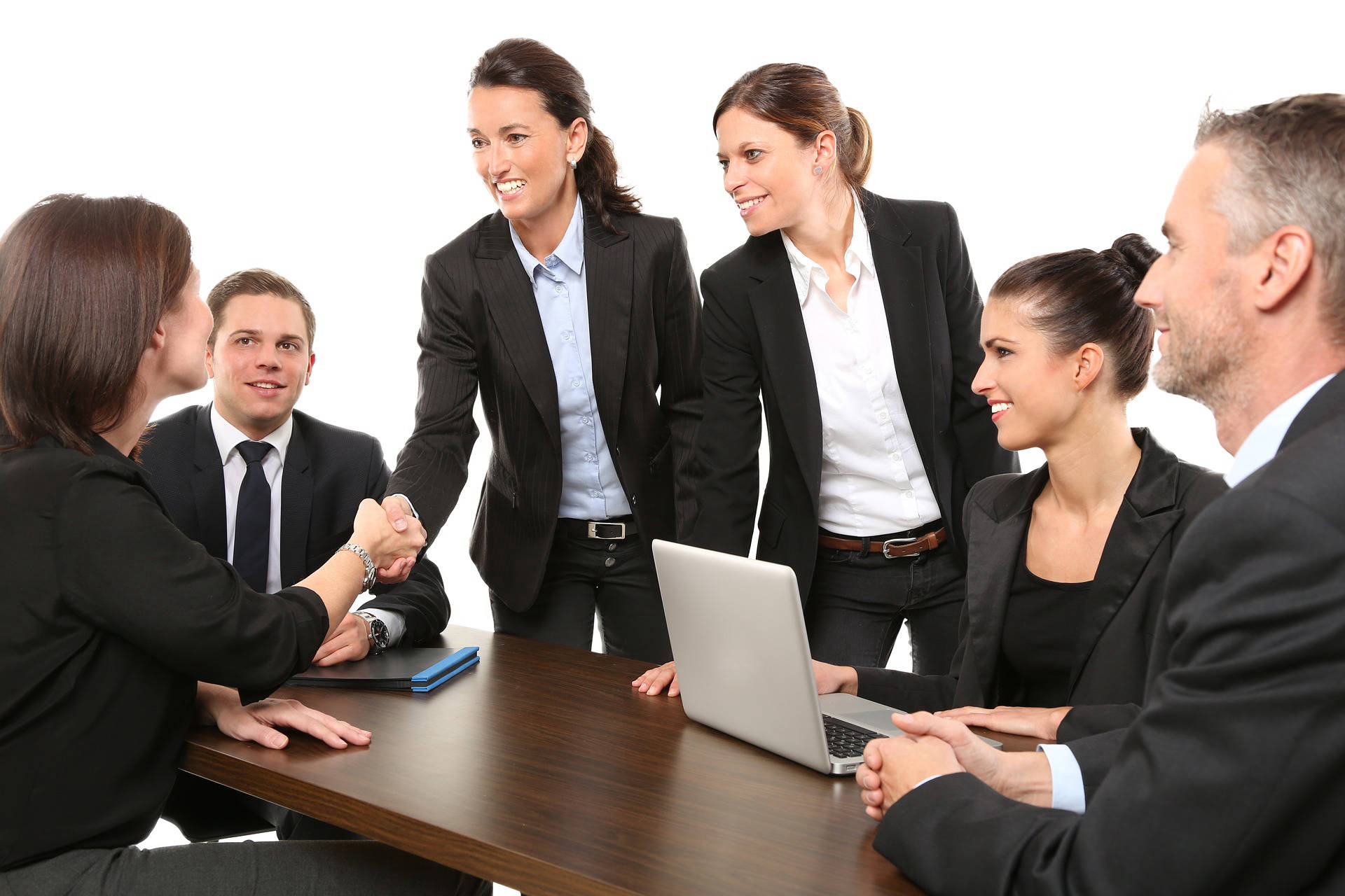 Six people in a meeting room with two people standing. One standing person is shaking hands with a seated person.